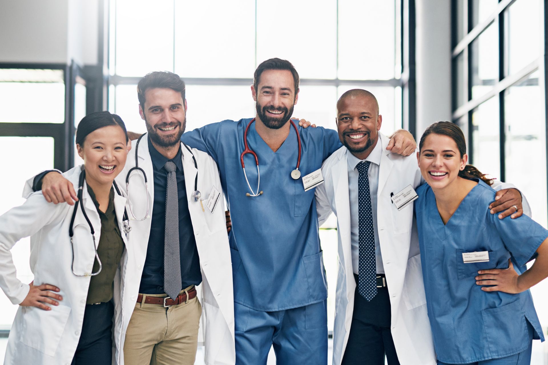 Porträt einer Gruppe von 5 Ärzten, die im Krankenhaus stehen, sich umarmen und in die Kamera lächeln
