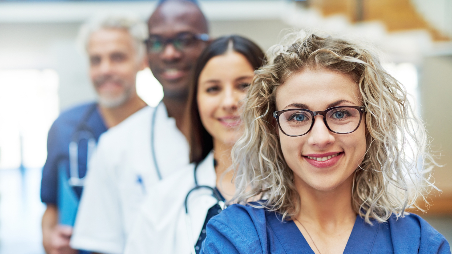 Fachkräfte 4 Fachkräfte Pflege/Medizin stehen seitlich versetzt, vordere Frau mit blonden lockigen haaren lächelt in die Kamera